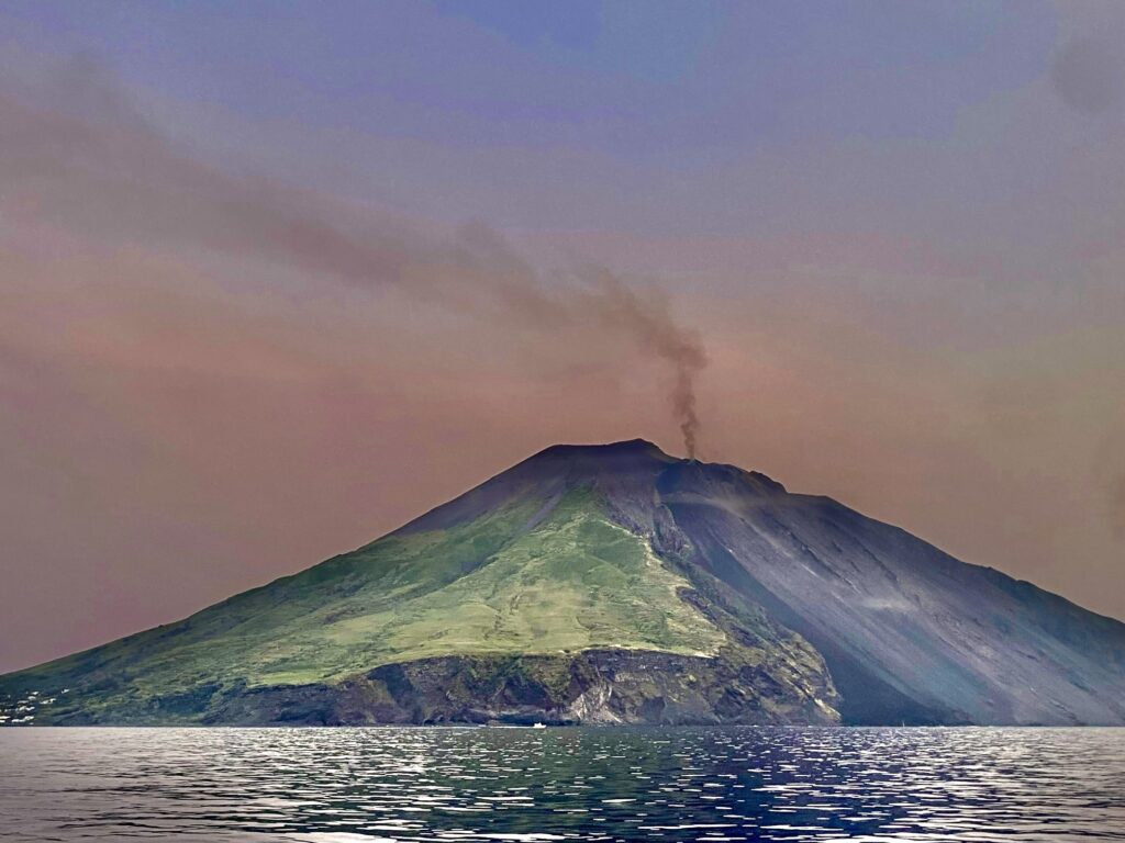 Volcano with green grass and black rock and smoke drifting above it.